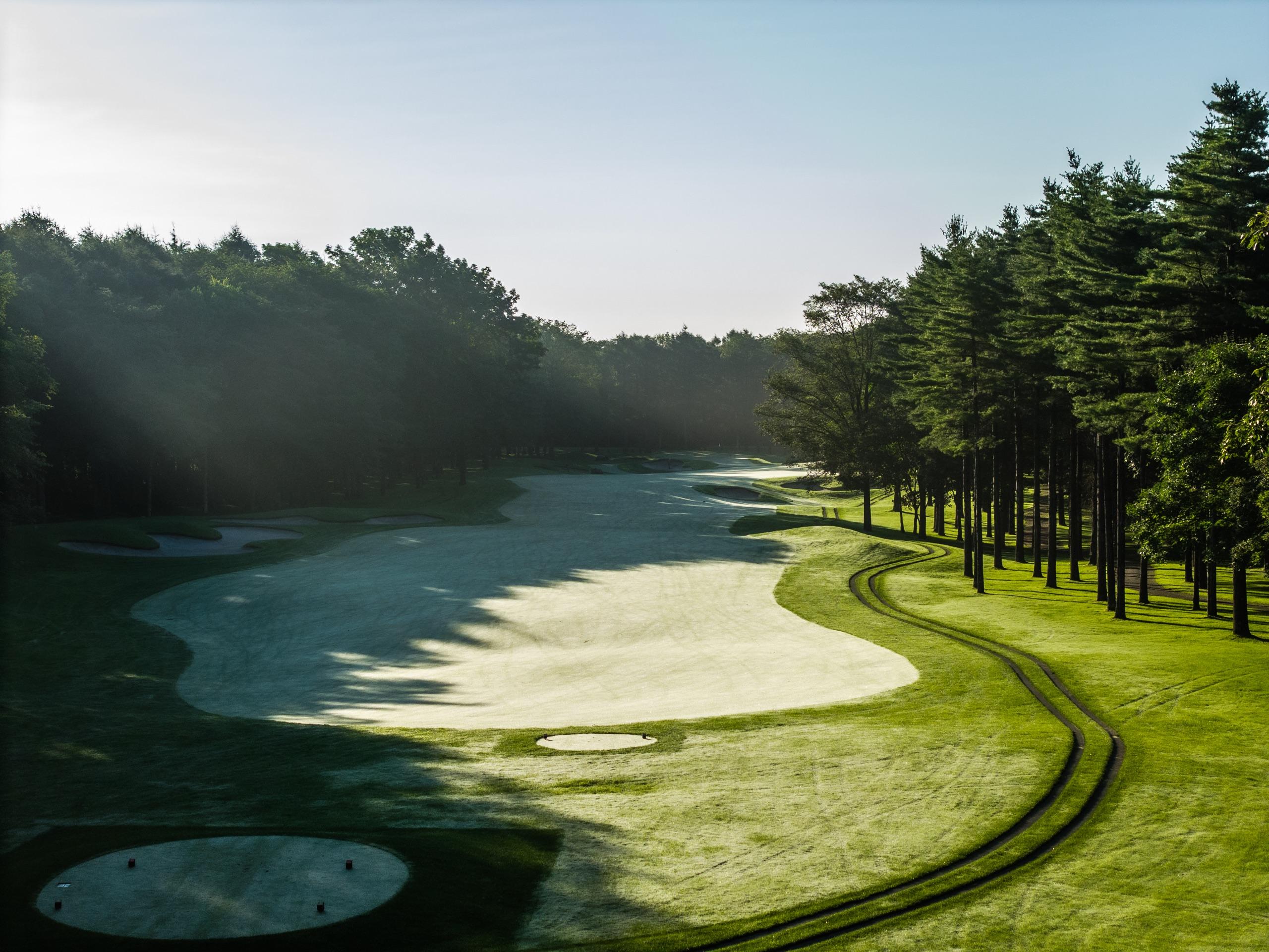 Morning shadows across a wide bentgrass fairway with bunkers left and a pine-lined cart path right