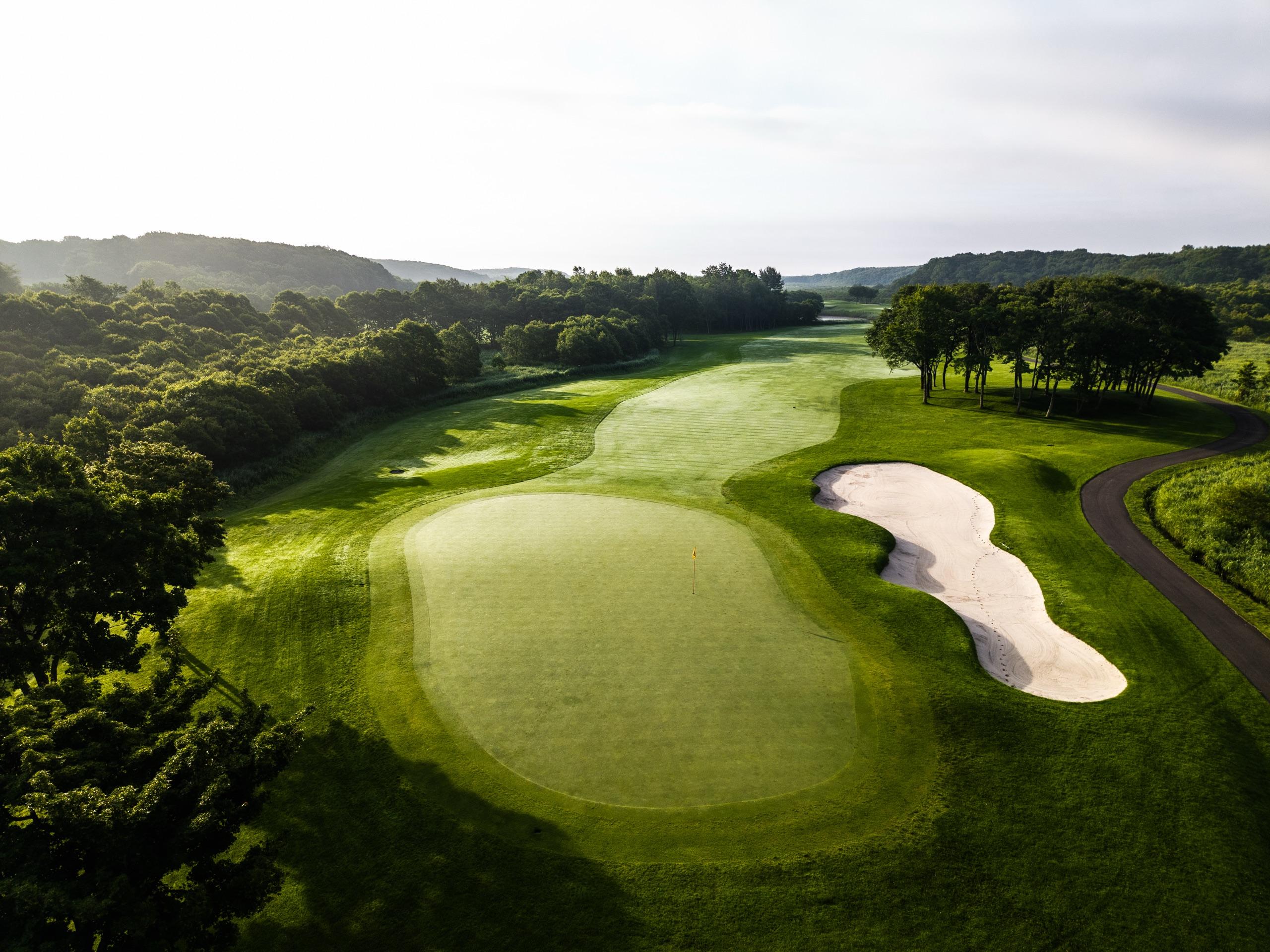 Morning light over a green with a long bunker on the right and a tree-lined fairway stretching into the distance
