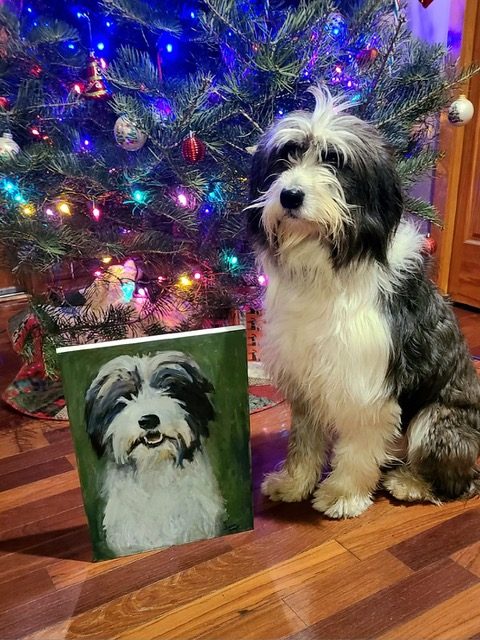 Otis, a black and white dog, sitting beside his painted portrait in front of a Christmas tree.