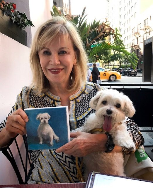 Val Sereno holding the painting of her small white dog, who sits beside her.