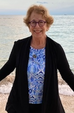 Jane Hart standing on a beach in a dark jacket and a blue patterned top, smiling.