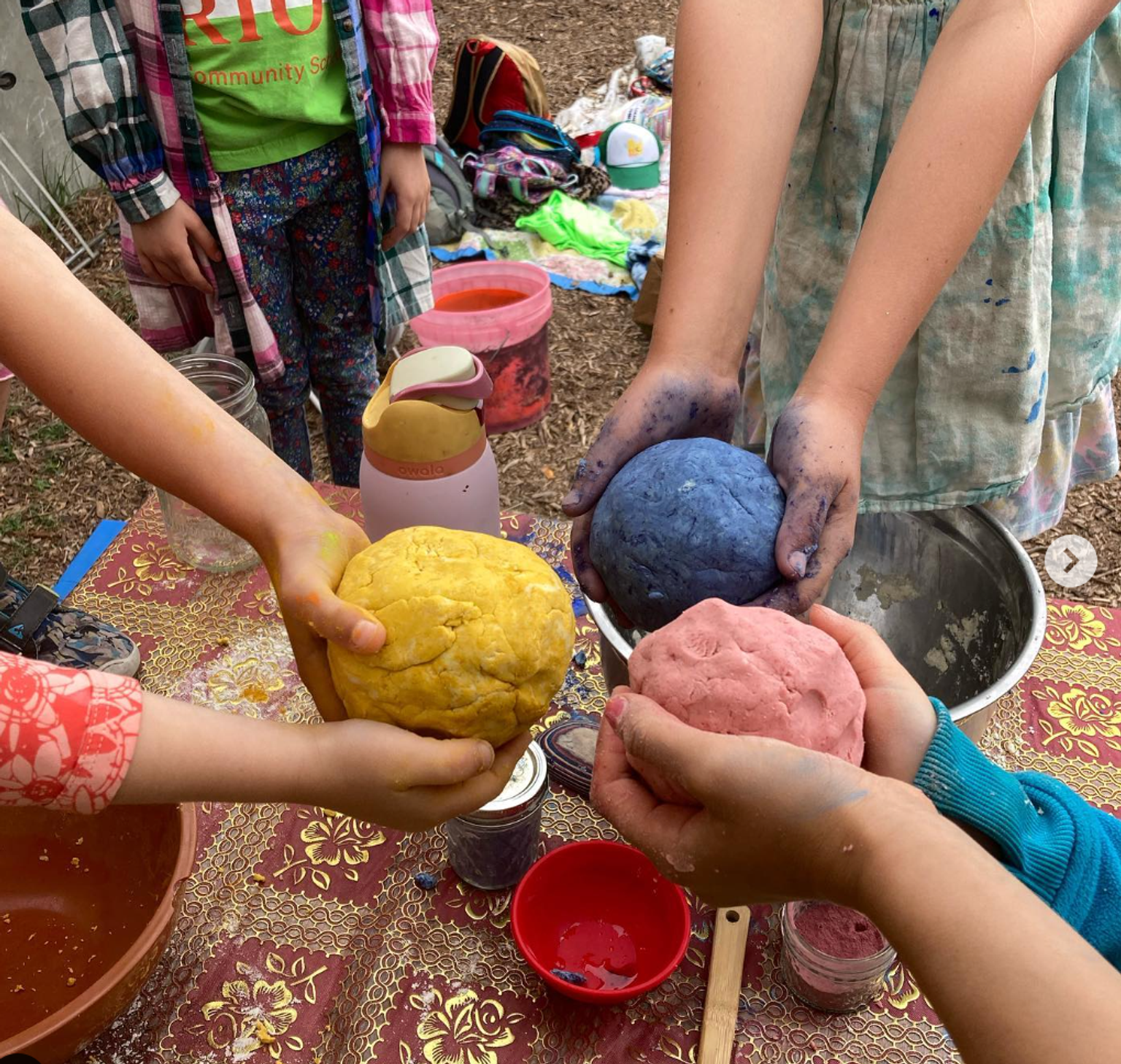 Children's hands hold pink, blue, and yellow tortilla doughs in a circle