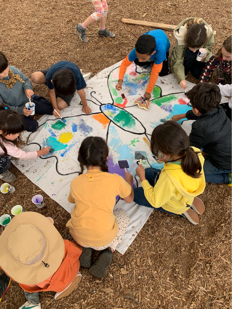 a group of children sitting around a large canvas banner with a butterfly outline, children are painting in the butterfly with many colors