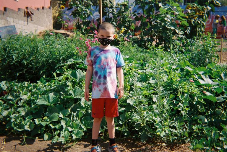 A child wearing a mask stands in front of garden greenery