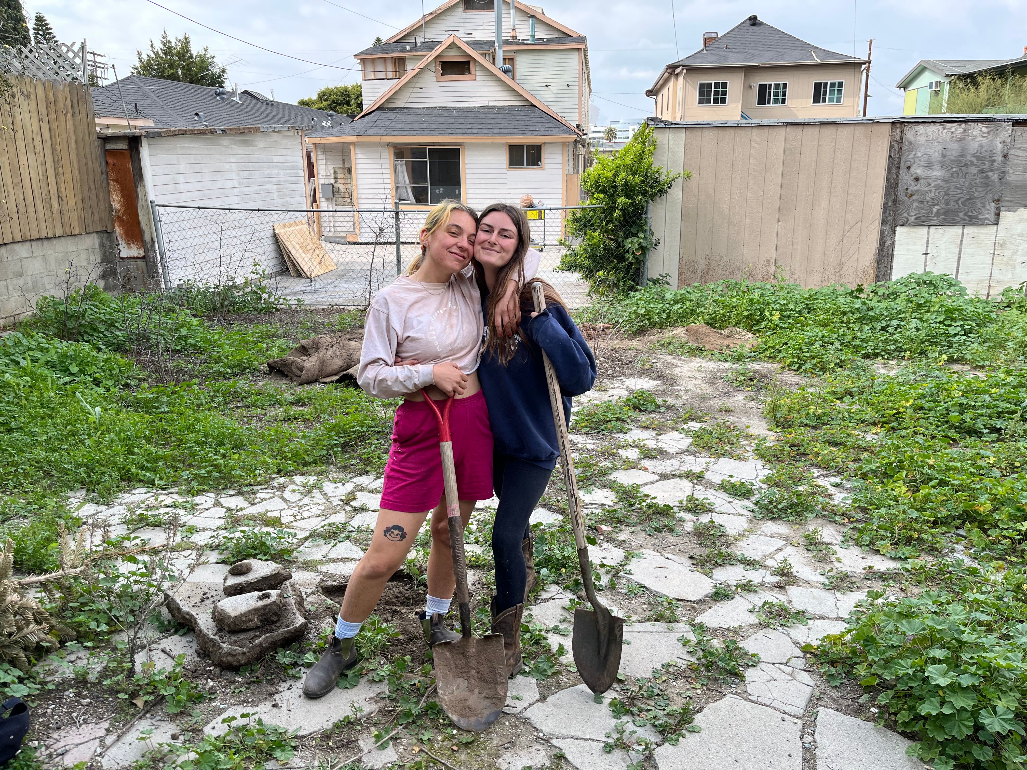 Two people standing in an empty lot with shovels, houses are in the background.