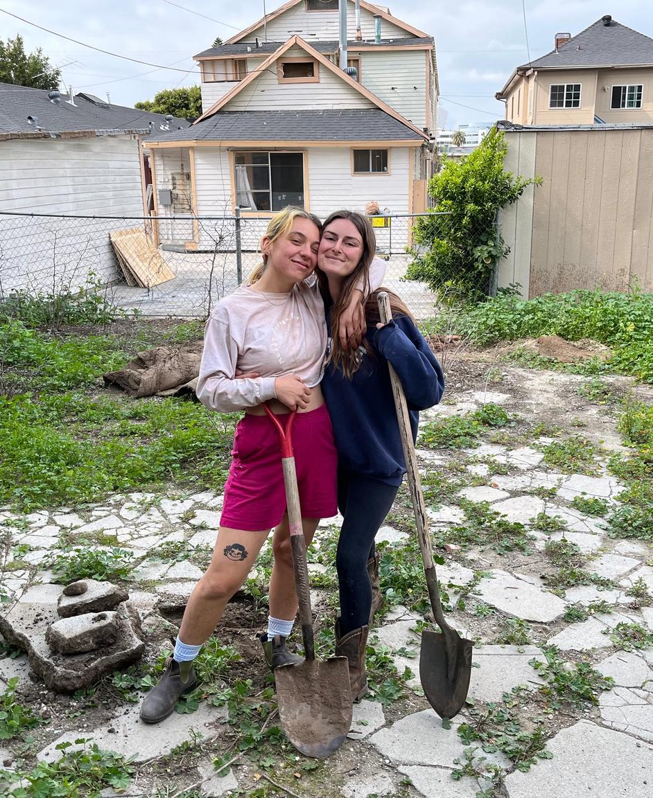 Two people standing in an empty lot with shovels, houses are in the background.