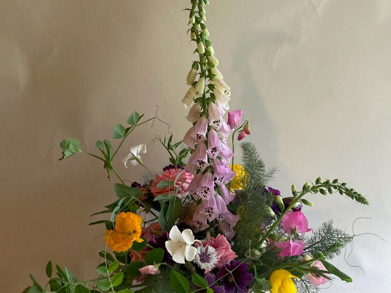 Fresh flower arrangement featuring a tall foxglove surrounded by pea tendrils and other small colorful flowers, against a light gray background