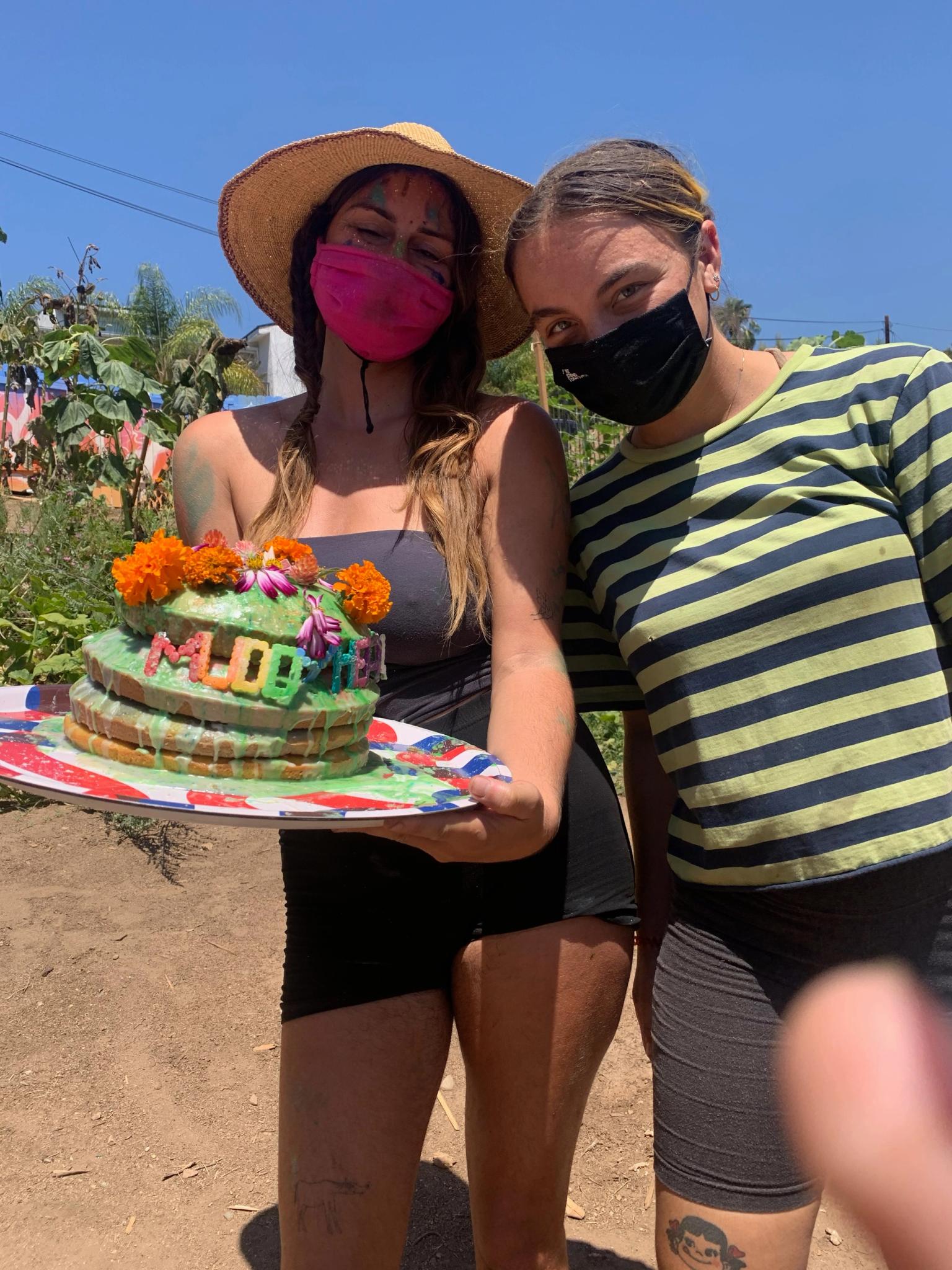 Two people wearing masks, person on the left is holding a tray with a cake with drippy green icing and the lettings Muddy Heaven