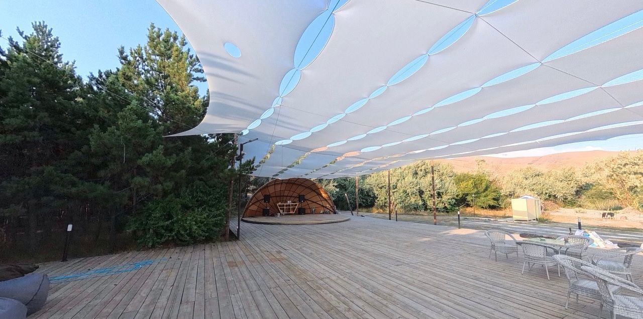 Alt text: Custom-built white tensile fabric shade structure covering a large outdoor wooden festival deck, with a stage, trees, and mountains in the background.