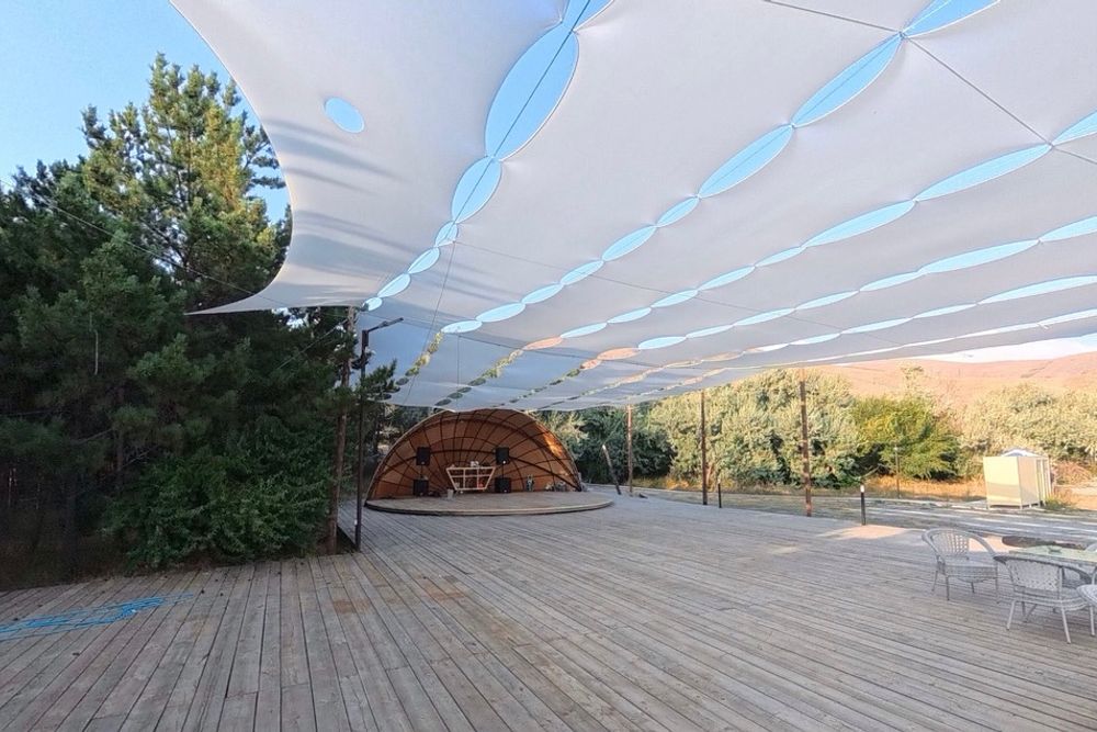 Alt text: Custom-built white tensile fabric shade structure covering a large outdoor wooden festival deck, with a stage, trees, and mountains in the background.