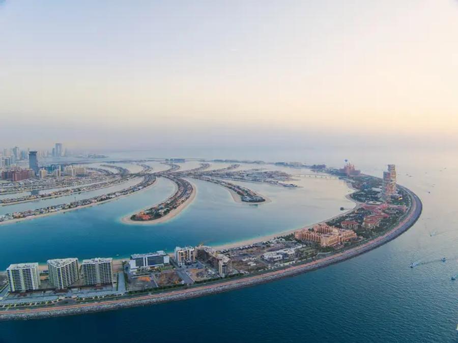 Aerial view of Dubai's Palm Jumeirah artificial island, its curved fronds lined with buildings, surrounded by turquoise sea under a hazy sunset sky.