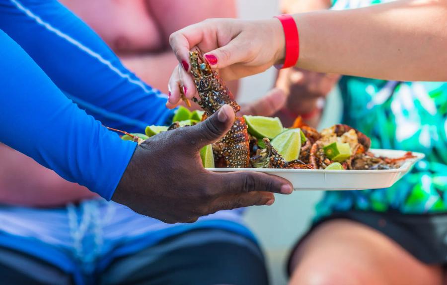 Close-up of one person holding a plate of cooked spiny lobster with lime wedges, while another person’s hand reaches in to pick up a lobster.