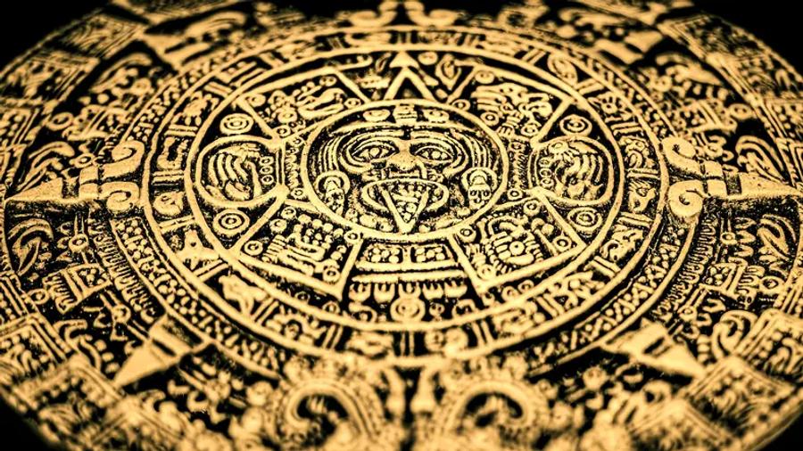 A gold-coloured circular carving with a face at its centre, surrounded by rings of intricate symbols and patterns, photographed at an angle against a black background.