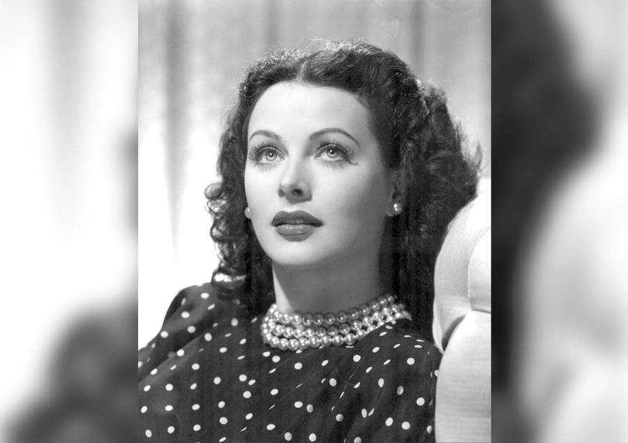 A black-and-white studio portrait of a woman with dark, curled hair, looking upwards. She wears a polka dot top and a thick, multi-strand pearl necklace.