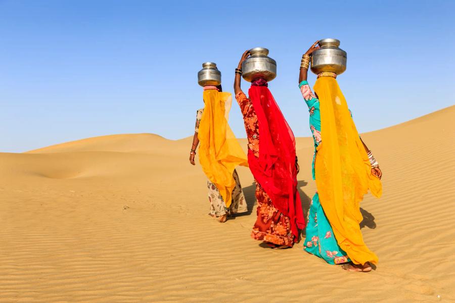 Three women in bright yellow, red and turquoise clothing, seen from behind, walking across sand dunes under a clear blue sky, each carrying a metal water pot on her head.