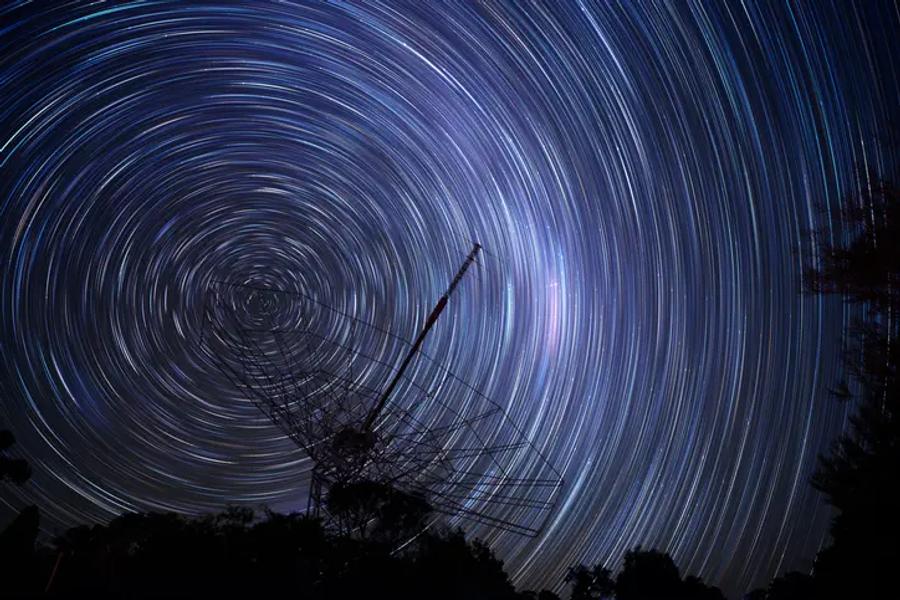 A long-exposure night photo of star trails swirling in circles around a point in the sky, above a large radio telescope dish and dark trees.