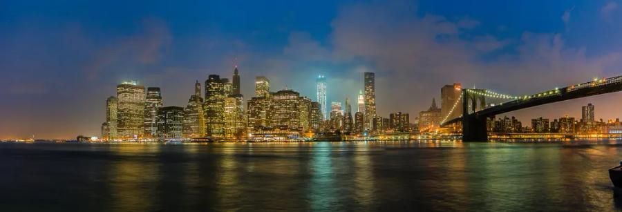 A city skyline of lit skyscrapers at night beside a bridge, reflected in dark water under a blue sky.