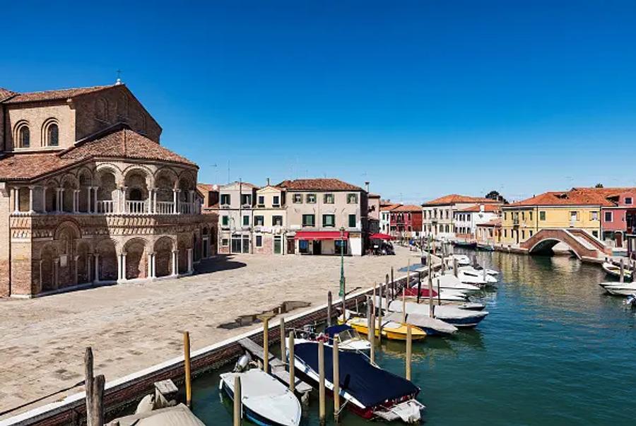 A sunny canal-side square in an Italian town, with an old brick church on the left, colourful houses along the water and small boats moored in front of a stone bridge.