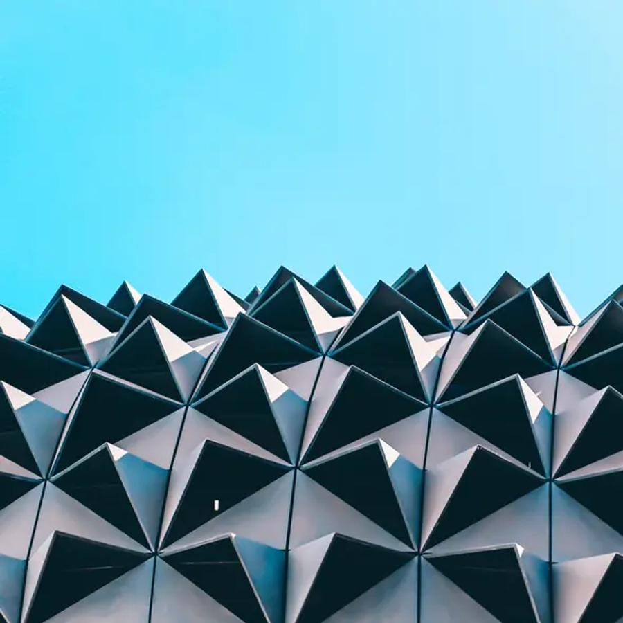 The top of a building facade covered in repeating angular panels shaped like small pyramids, in grey and white with deep shadows, against a clear blue sky.