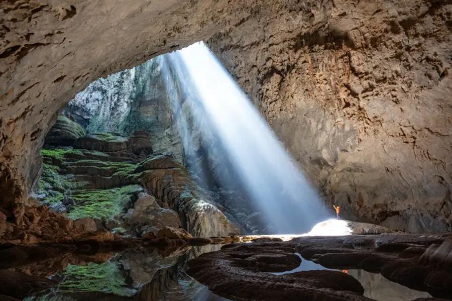 A huge cave with a beam of sunlight pouring through an opening in the roof onto rocky ground, with green moss on the rocks and a tiny person standing in the light.