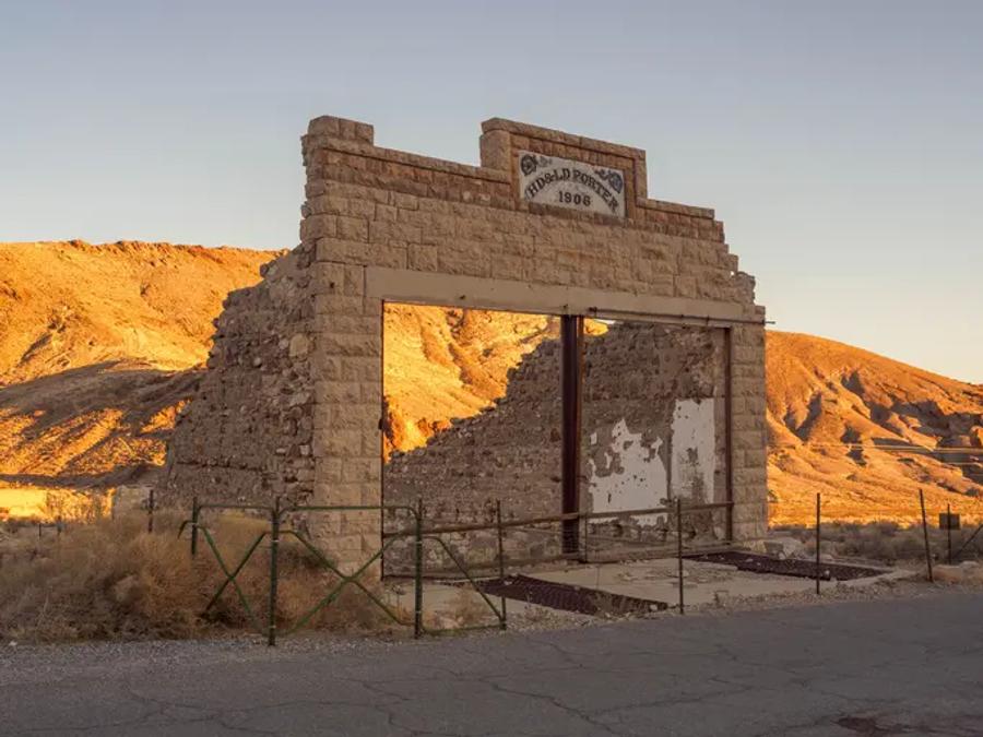 The ruined stone front of an old building standing in the desert, its doorway open to the sky with no roof behind it. A sign above reads "Porter" and "1906", and dry golden hills rise in the background in low evening light.