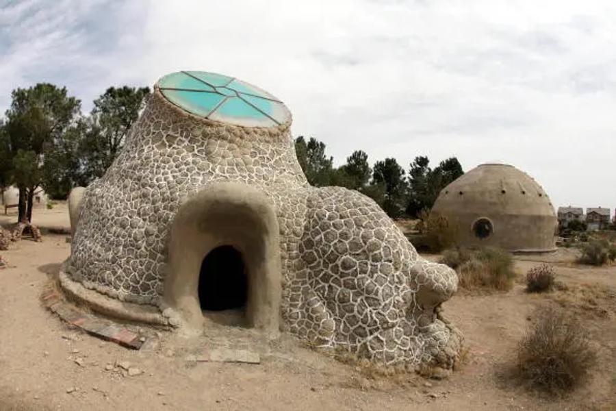 A dome-shaped earth building covered in a pattern of stones, with a round arched doorway and a skylight of green and blue panels on top, and a second smaller dome behind it in a desert setting.