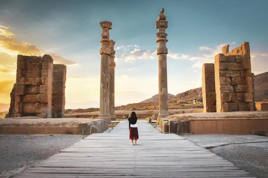 A visitor stands on a wooden walkway facing the tall stone columns and gateway ruins of Persepolis, with golden sunset light behind them.