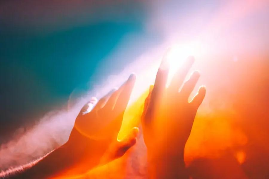 hands reach up to a blue, pink and orange sky with bright sunlight