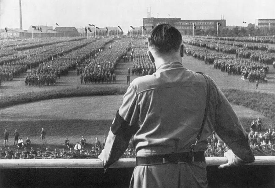 A black-and-white photograph of a man seen from behind, in a uniform with an armband and shoulder strap, standing at a stone balustrade. He looks out over huge ranks of uniformed men in formation across a large field, with flags and buildings in the distance.