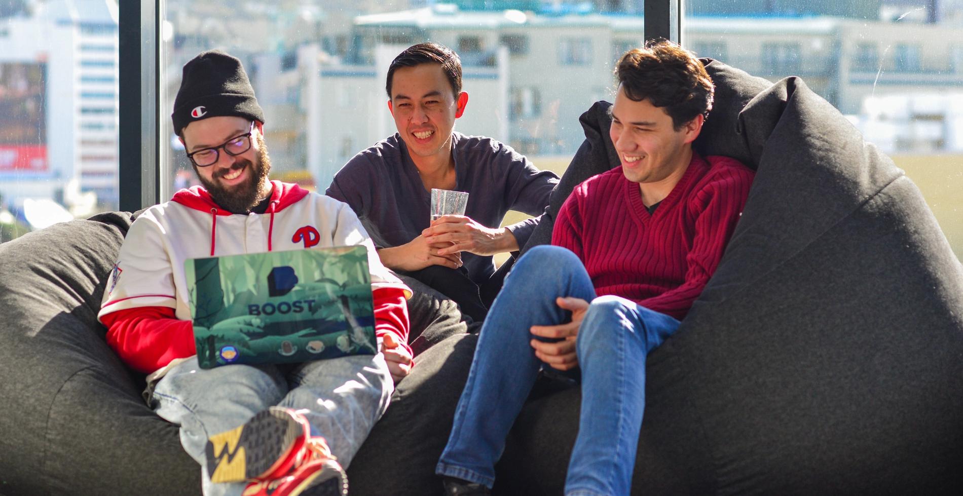 The image shows three young men sitting on a beanbag and looking at a laptop.
