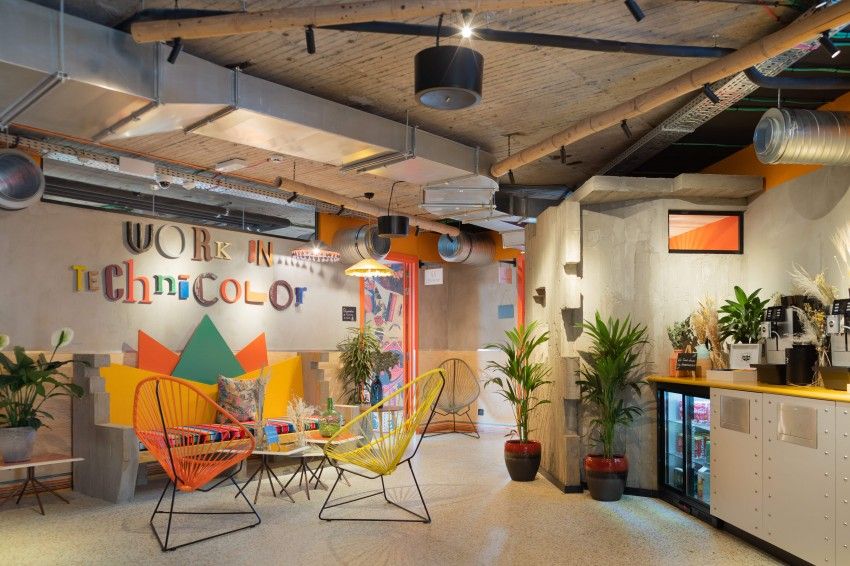 Een kleurrijke lounge met oranje en gele Acapulco-stoelen, planten en het muuropschrift "Work in Technicolor", naast een drankenbar.