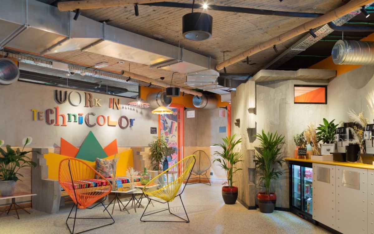 A colourful lounge with orange and yellow Acapulco chairs, plants and the wall lettering "Work in Technicolor", next to a drinks counter.