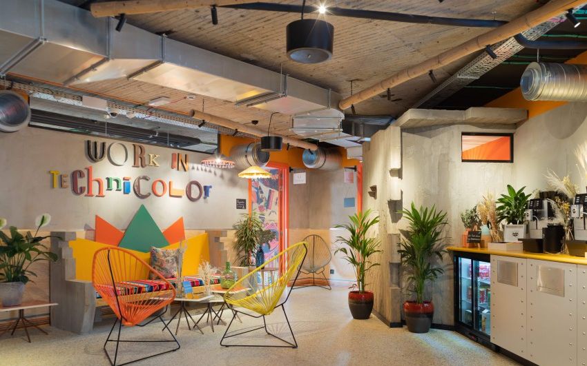 Salon coloré avec fauteuils Acapulco orange et jaune, plantes et lettrage mural « Work in Technicolor », près d'un comptoir de boissons.