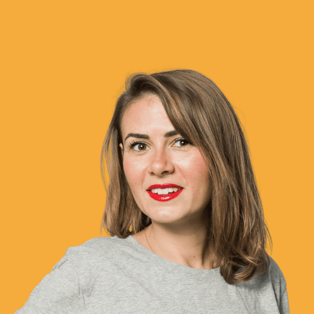 Julie Gamundi, glimlachend, met halflang haar en een grijs T-shirt, tegen een gele achtergrond.