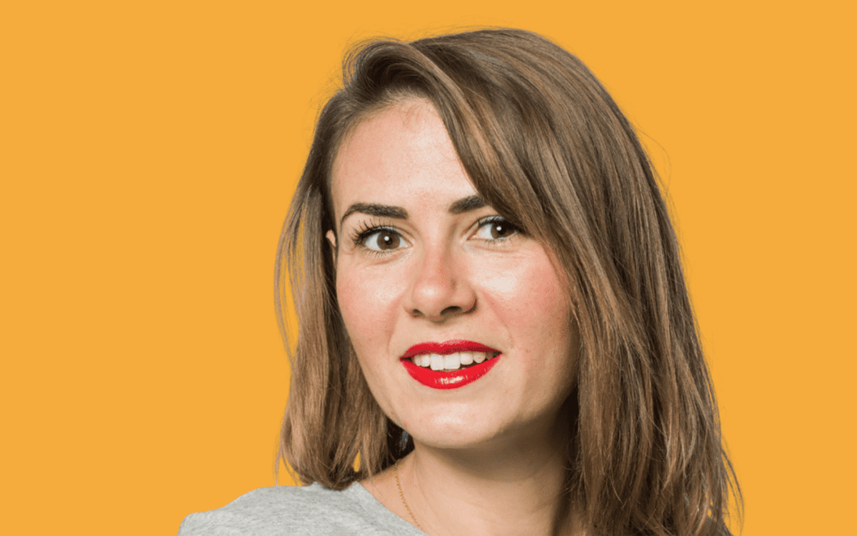 Julie Gamundi, glimlachend, met halflang haar en een grijs T-shirt, tegen een gele achtergrond.