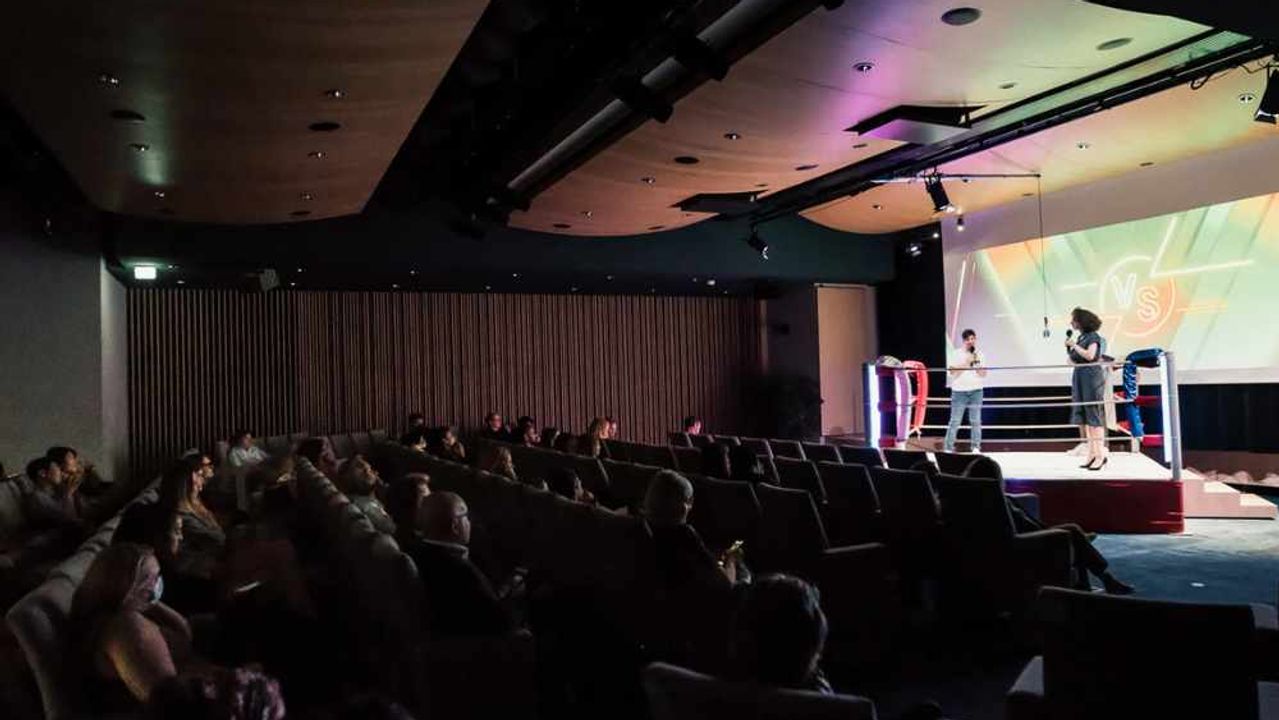 In a dimly lit auditorium, an audience seated in armchairs watches two speakers with microphones standing in a lit boxing ring in front of a giant screen.