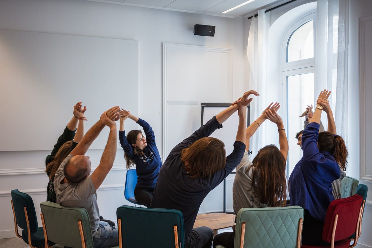 Een groep deelnemers strekt zich met geheven armen uit tijdens een welzijnssessie in een lichte vergaderzaal.