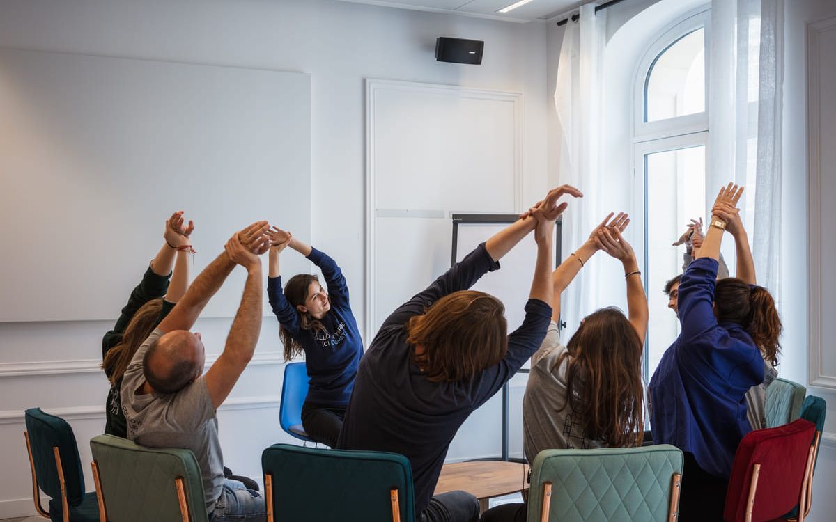 Een groep deelnemers strekt zich met geheven armen uit tijdens een welzijnssessie in een lichte vergaderzaal.