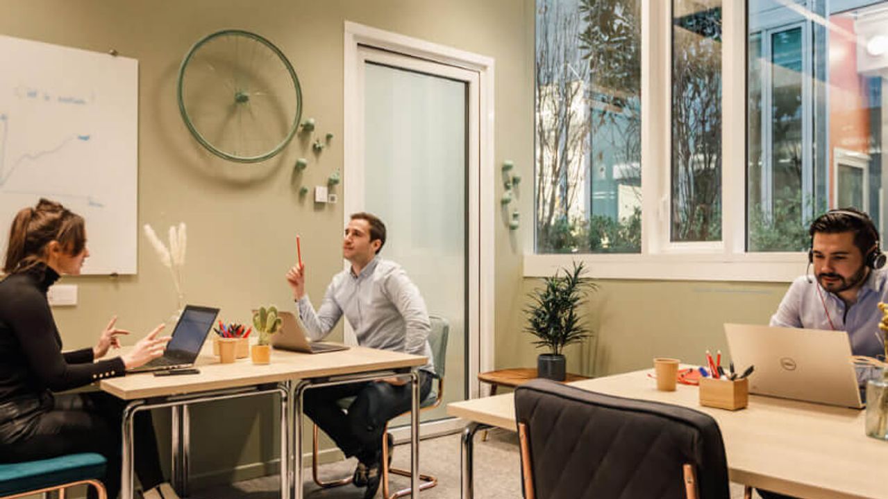 Three people working on laptops in a light green room decorated with a bicycle wheel and opening onto large windows.