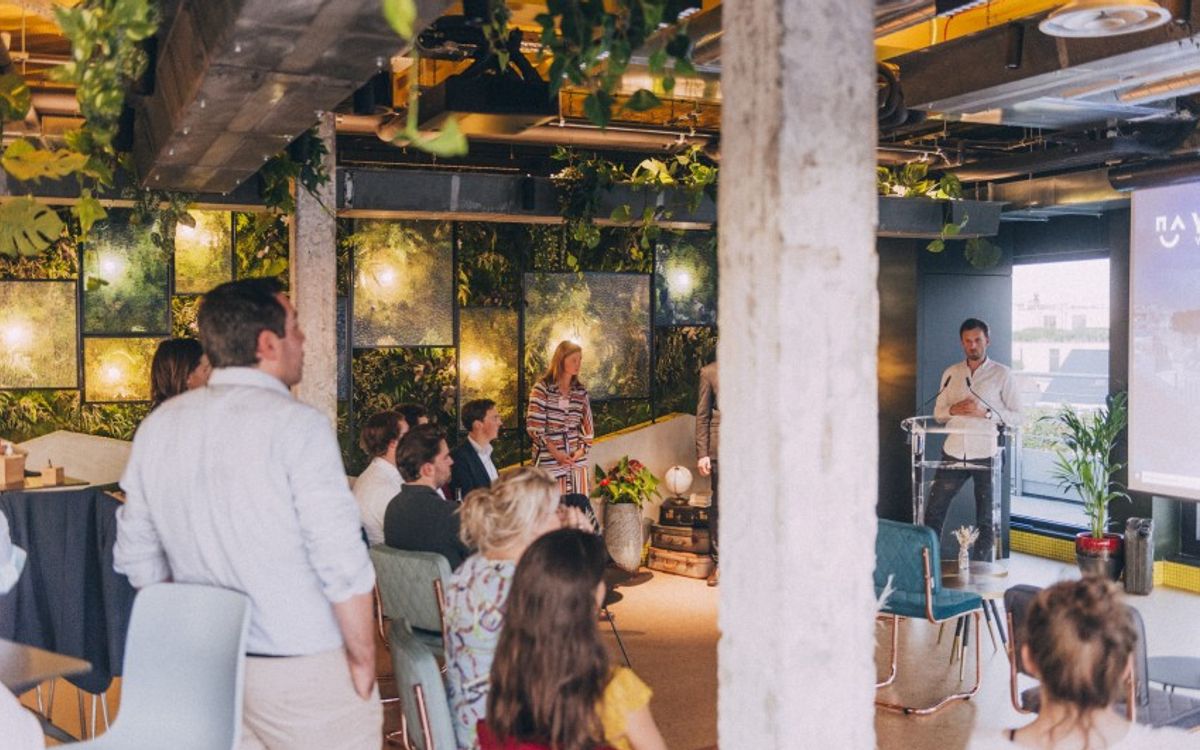 A seated audience listens to a speaker at a lectern in an event space with plant-covered walls and a view over the city.