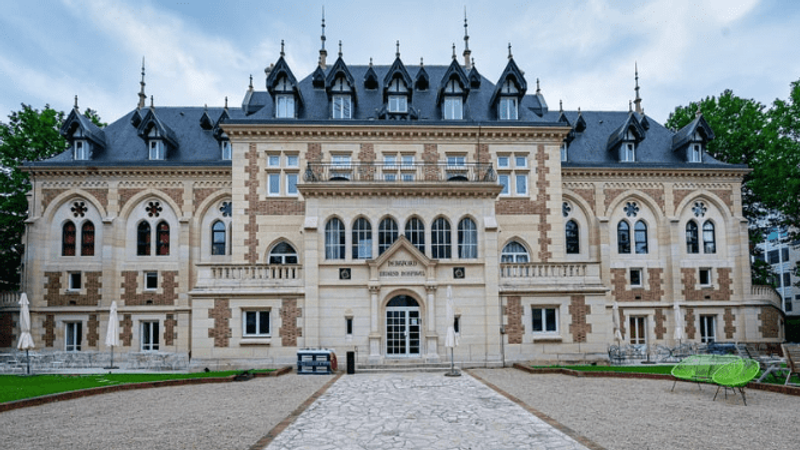 The neo-Gothic façade of the Levallois château, topped with slate roofs, at the end of a gravel driveway lined with lawn.