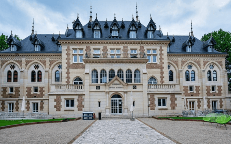 The neo-Gothic façade of the Levallois château, topped with slate roofs, at the end of a gravel driveway lined with lawn.