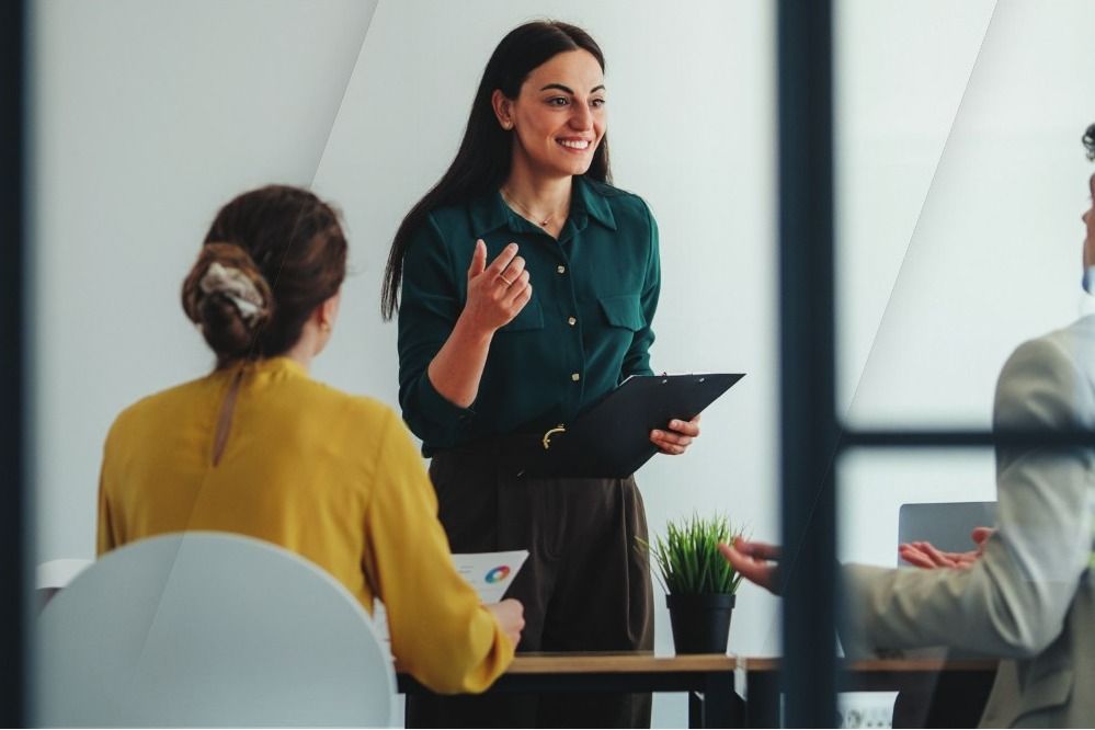 A smiling manager stands and talks with her team around a meeting table, seen through a glass partition.