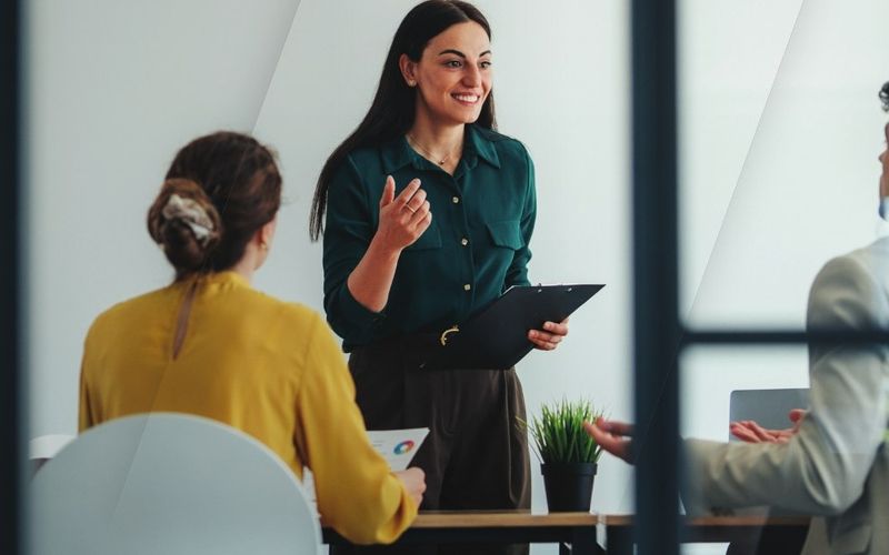 A smiling manager stands and talks with her team around a meeting table, seen through a glass partition.
