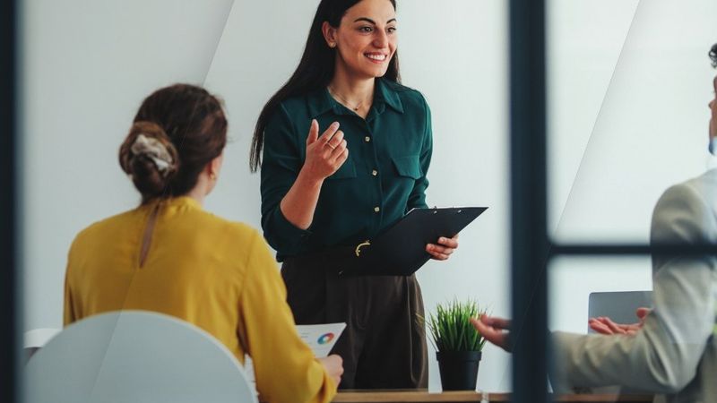 Una mánager de pie, sonriente, conversa con su equipo alrededor de una mesa de reuniones, vista a través de una mampara de cristal.