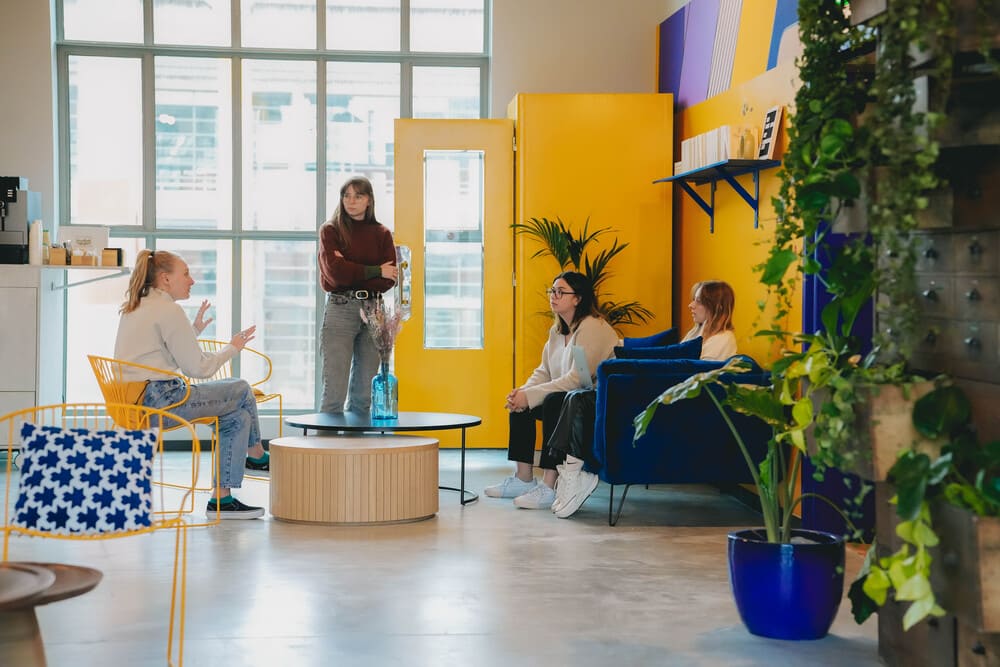 Four people talking in a lounge with yellow doors and a blue sofa, in front of tall glass windows and a wall of plants.