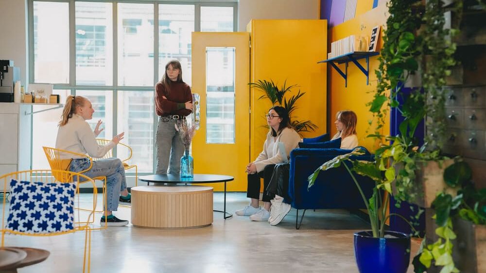 Quatre personnes échangent dans un salon aux portes jaunes et au canapé bleu, devant de grandes verrières et un mur végétalisé.