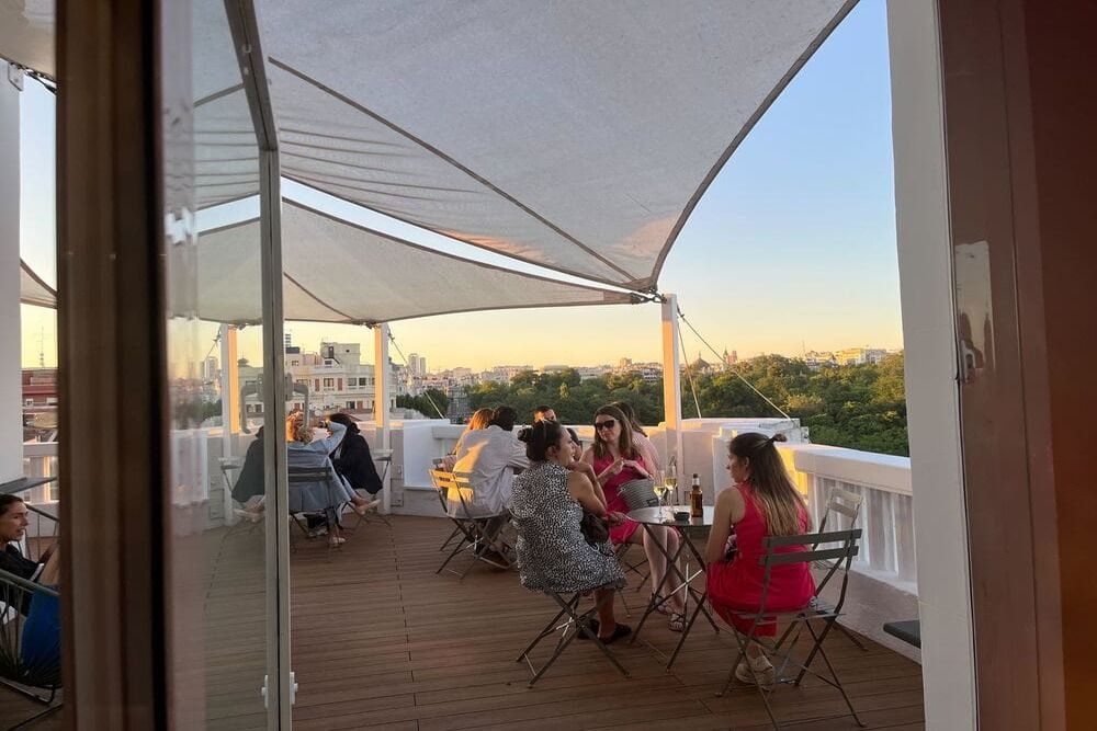 Groepjes aan tafeltjes op een houten terras onder witte schaduwdoeken bij zonsondergang, met zicht op de bomen van het Retiro-park en de daken van Madrid.