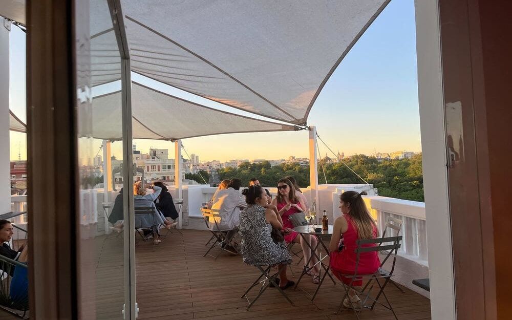 Des groupes attablés sur une terrasse en bois abritée par des voiles blanches, au coucher du soleil, face aux arbres du Retiro et aux toits de Madrid.