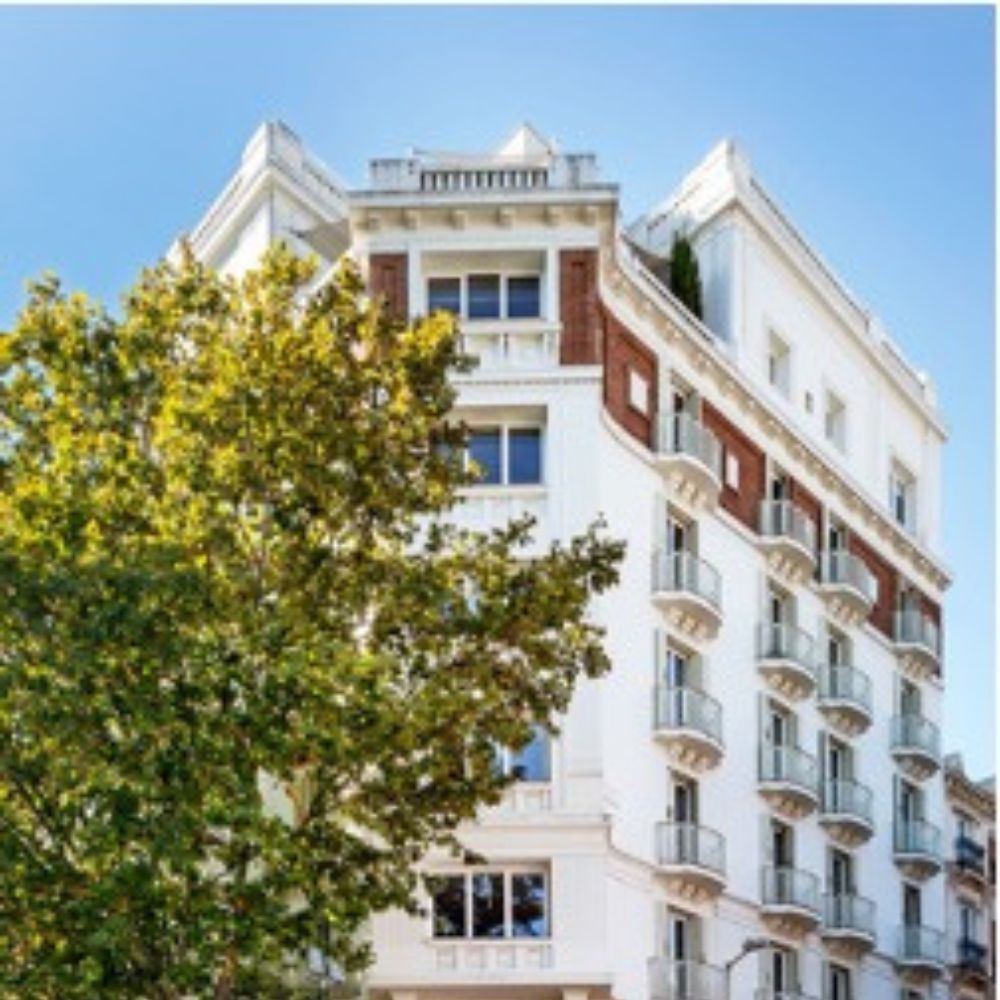 La façade blanche d'un immeuble madrilène aux balcons ouvragés, partiellement masquée par un arbre, sous un ciel bleu.